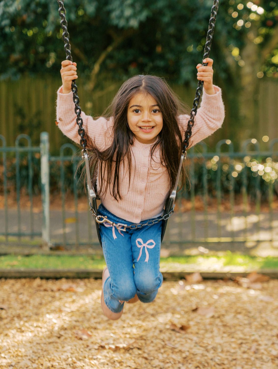 A smiling young girl with long dark hair swings on a playground swing, wearing a pink top and blue jeans.