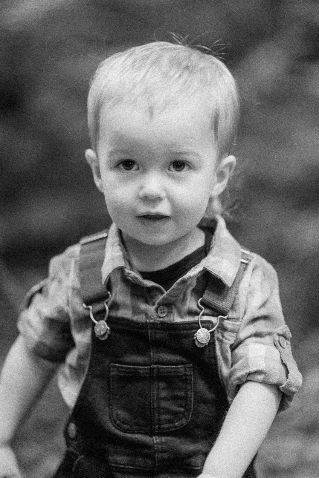 Black and white portrait of a toddler boy wearing denim overalls and plaid shirt outdoors.