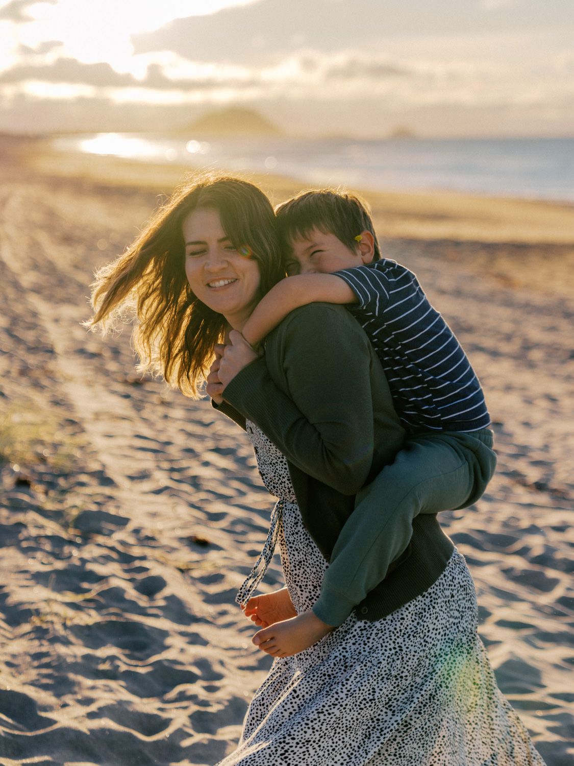 A woman gives a young boy a piggyback ride on a sandy beach at golden sunset, both smiling joyfully.