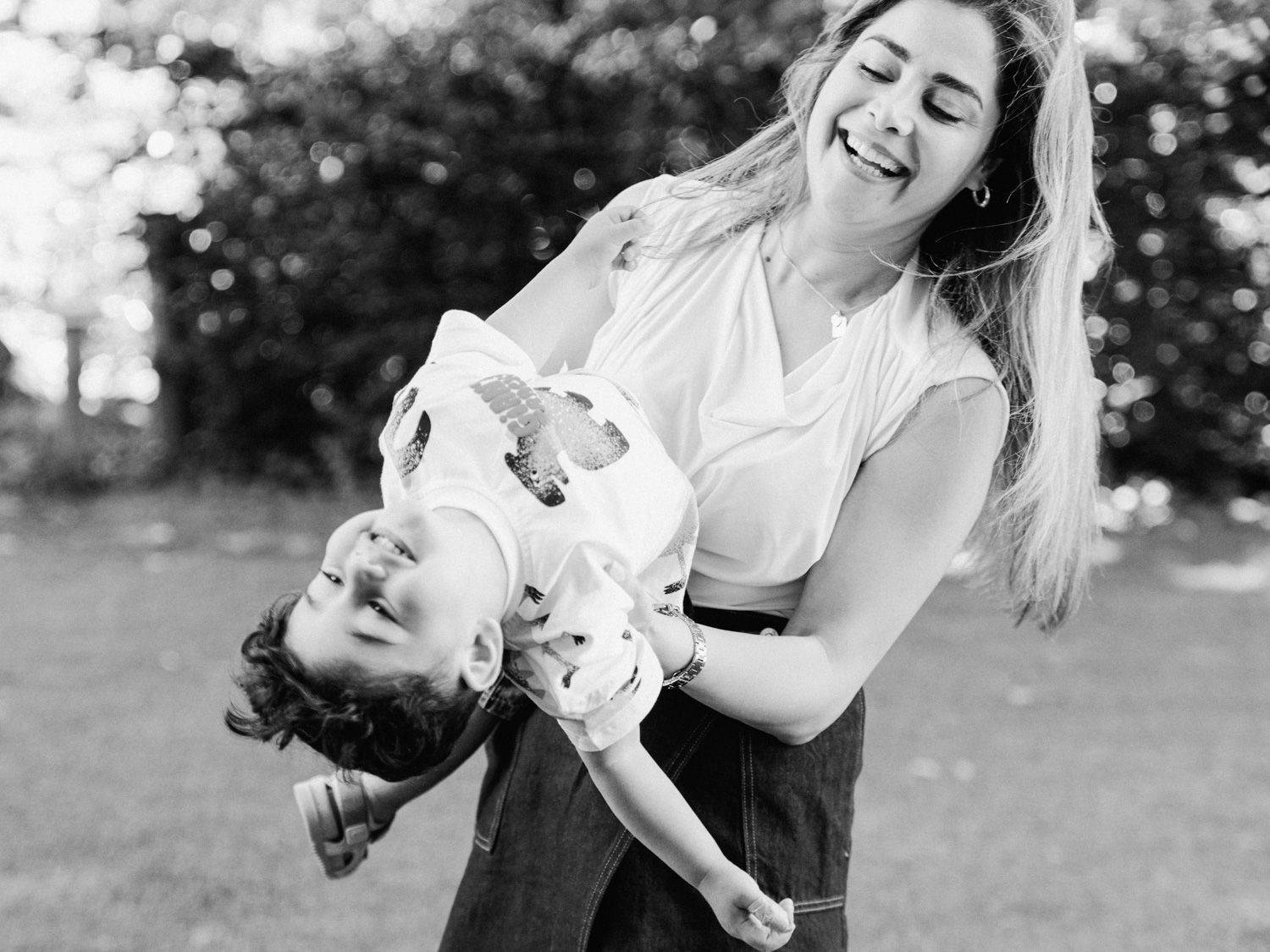 A laughing woman playfully holds a young child upside down outdoors in a lush green park, black and white photo.