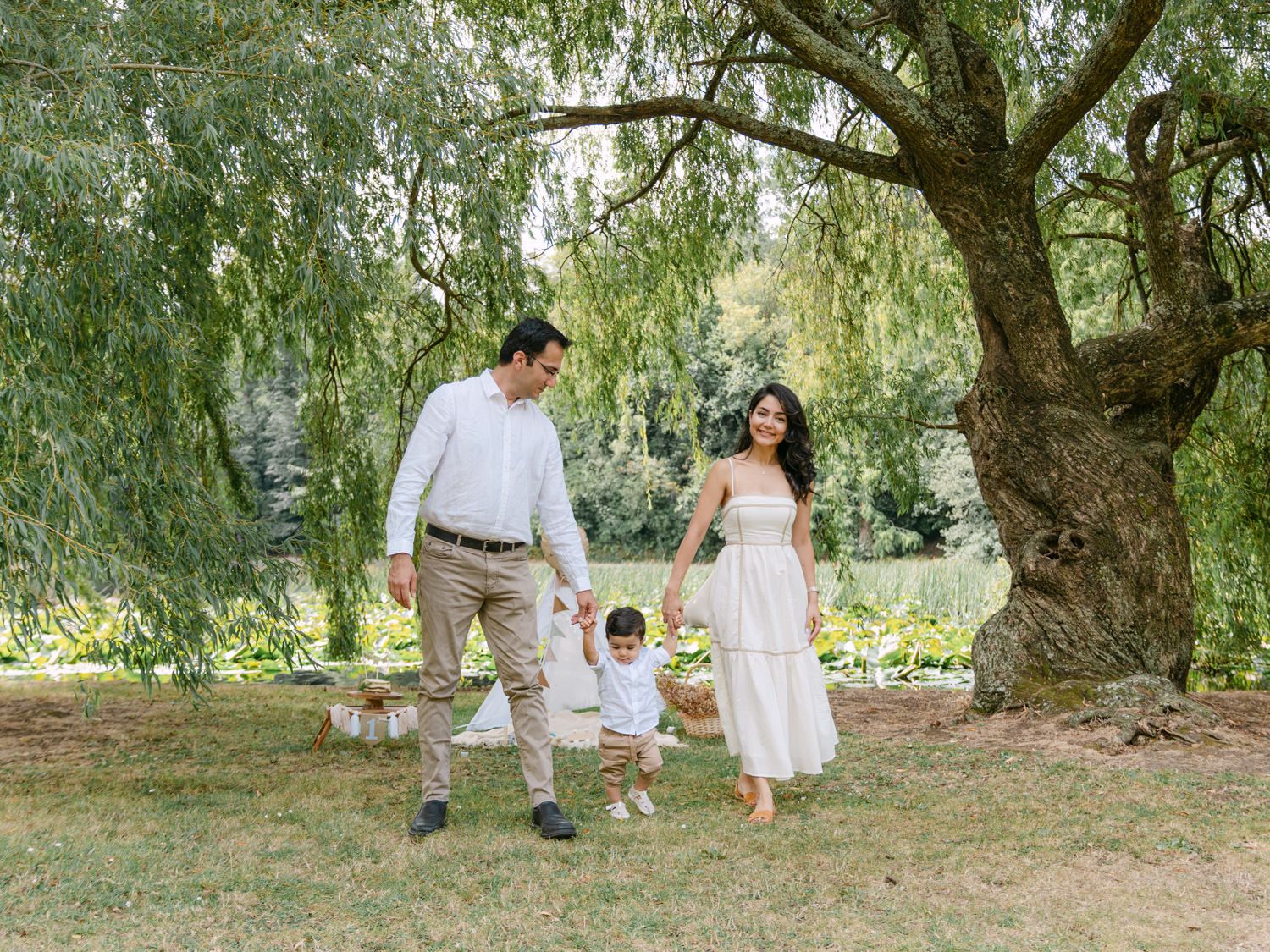 Family of three walking under a willow tree in a lush green park, parents swinging toddler between them.