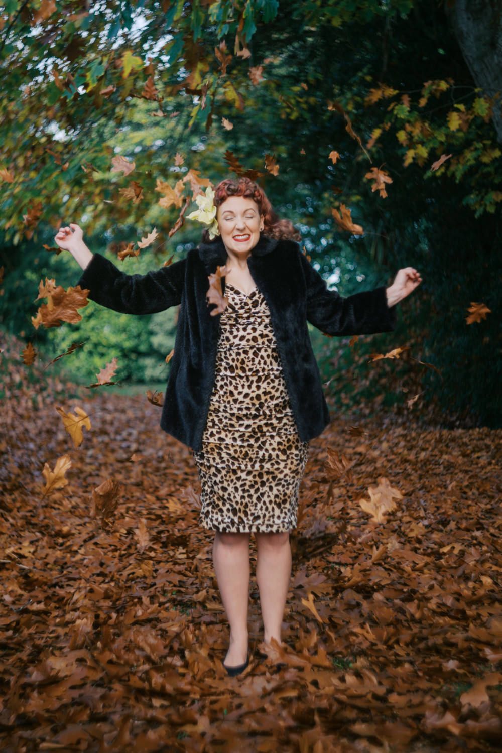 Woman in leopard dress and fur coat joyfully tossing autumn leaves in a forest setting.