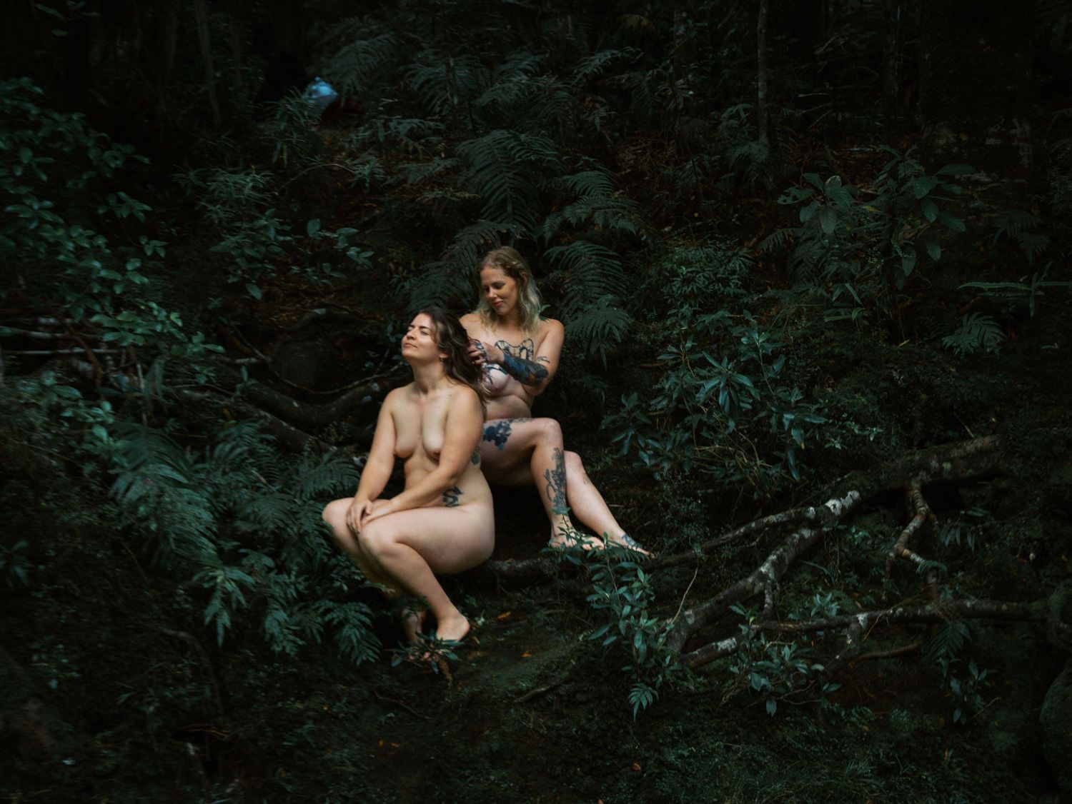 Two women sitting among lush green ferns and foliage in a dark forest setting.