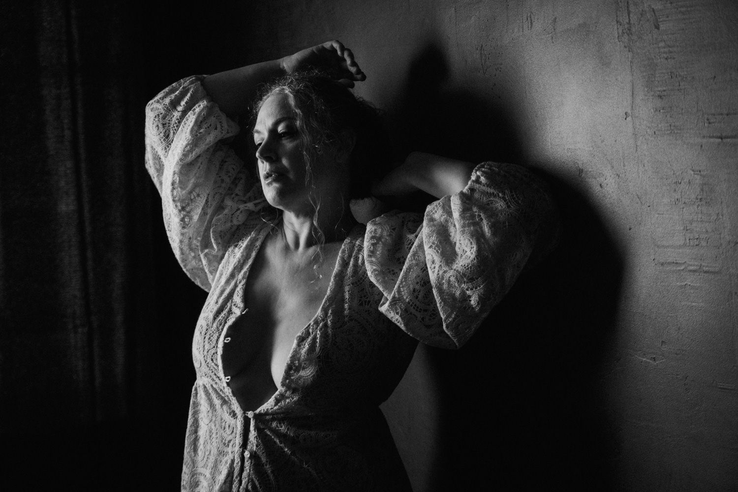 Black and white artistic boudoir portrait of woman in lace robe against dark wall.