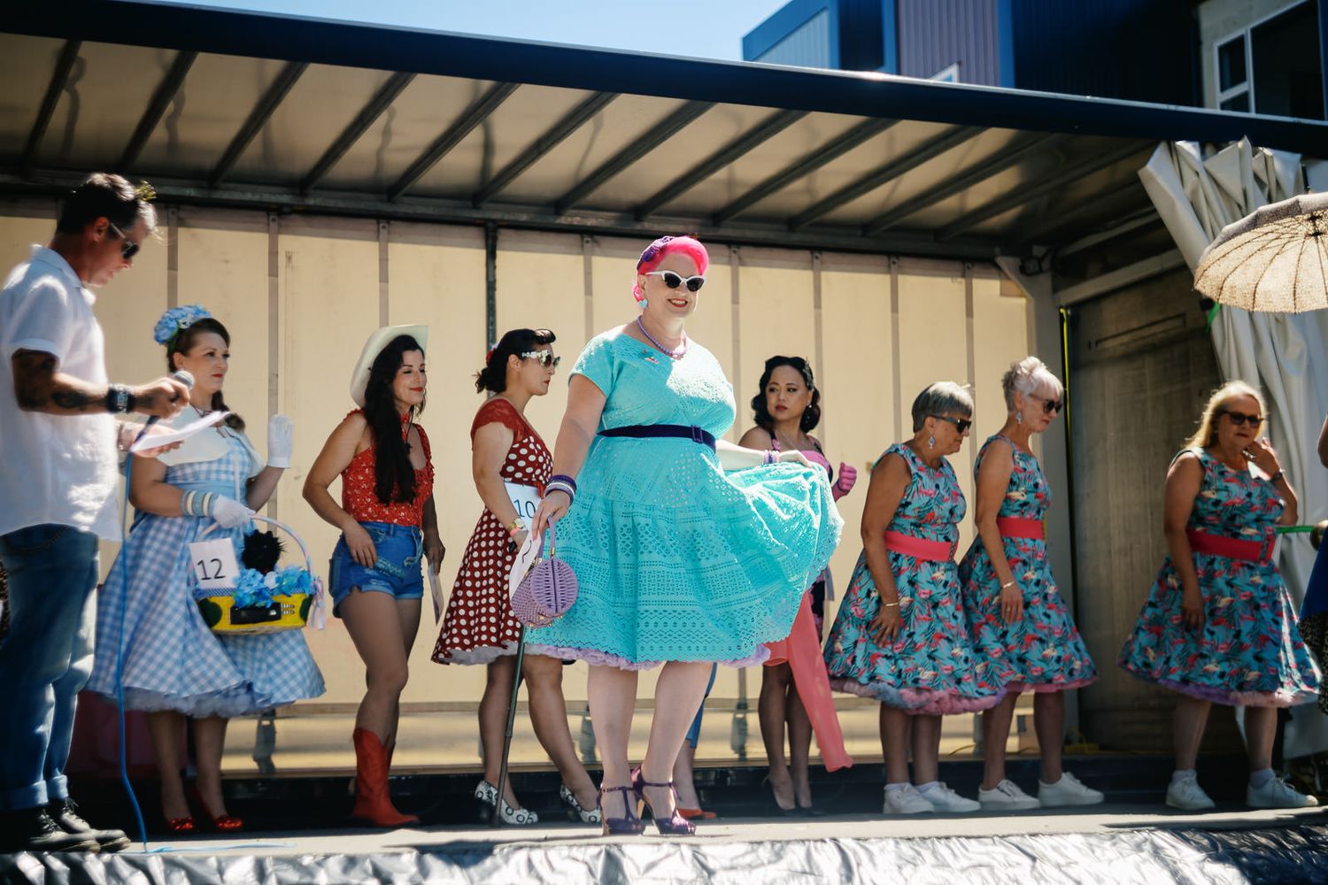 Women in colorful 1950s-style polka dot dresses line up on a stage at a vintage fashion contest outdoors.