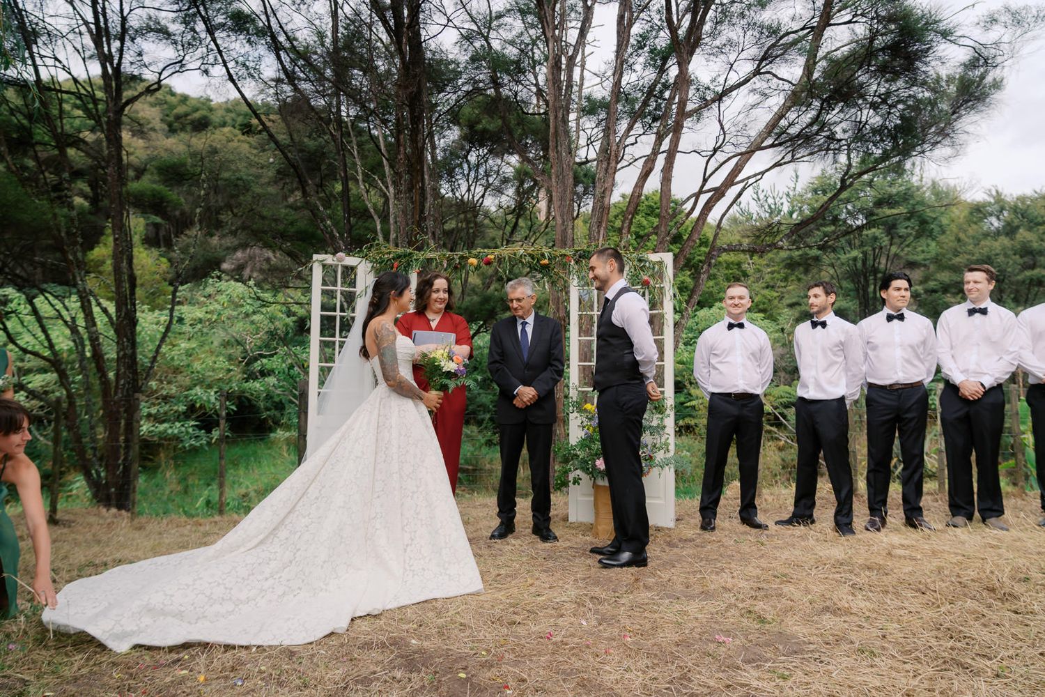 Outdoor wedding ceremony with bride in white gown, groom, officiant, and groomsmen standing among trees.