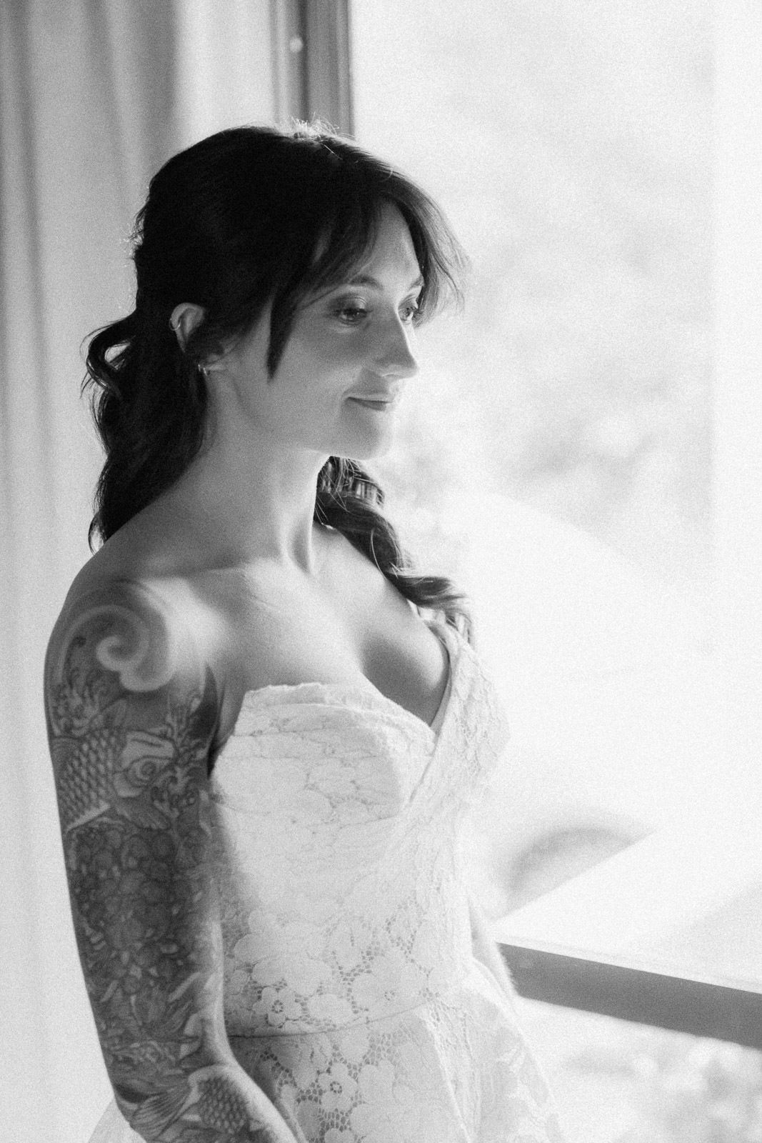 Black and white photo of a tattooed bride in a strapless lace wedding gown smiling softly by a bright window.