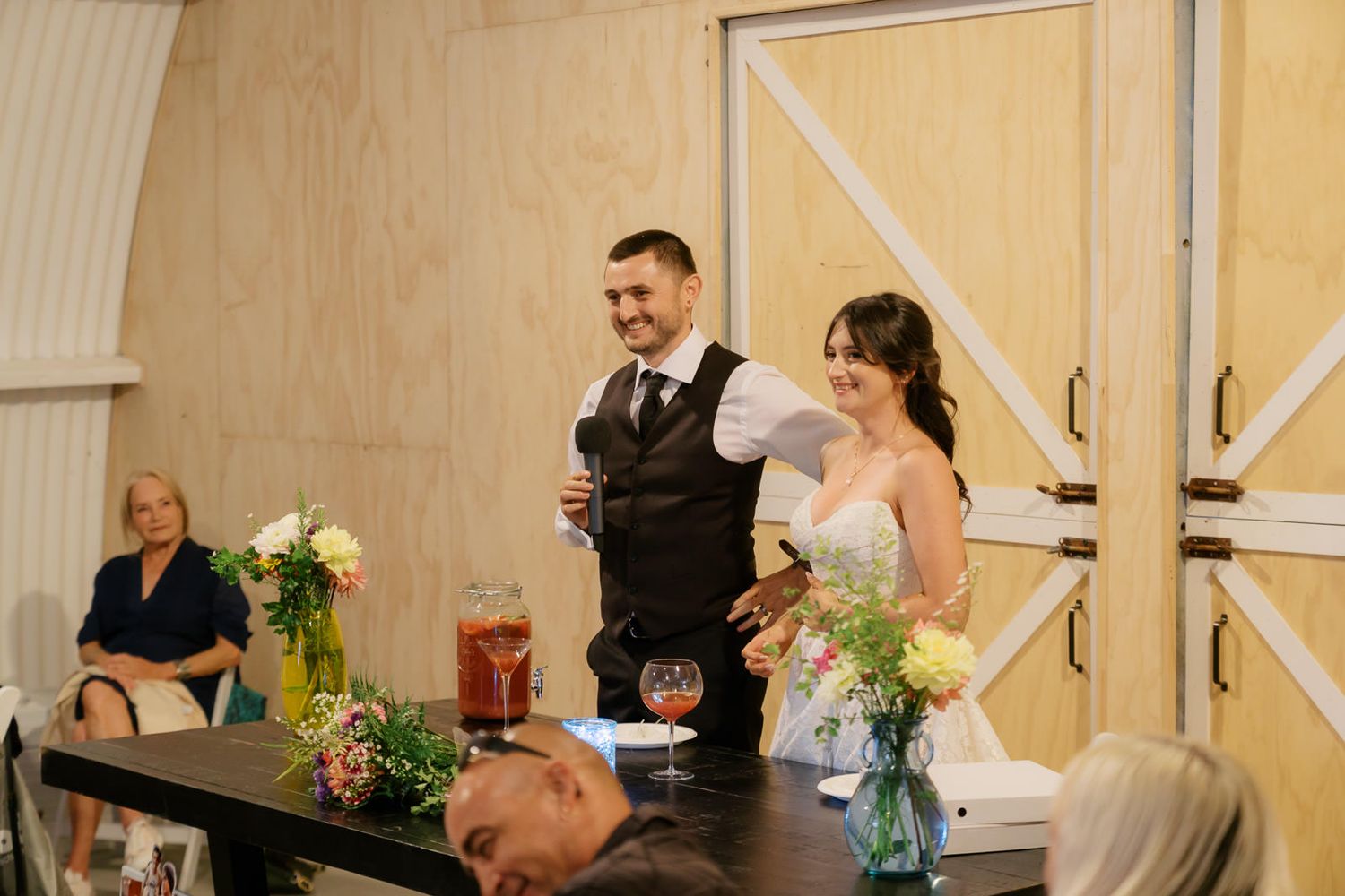 Bride and groom smiling together at wedding reception, holding wine glasses near floral arrangements in barn venue.