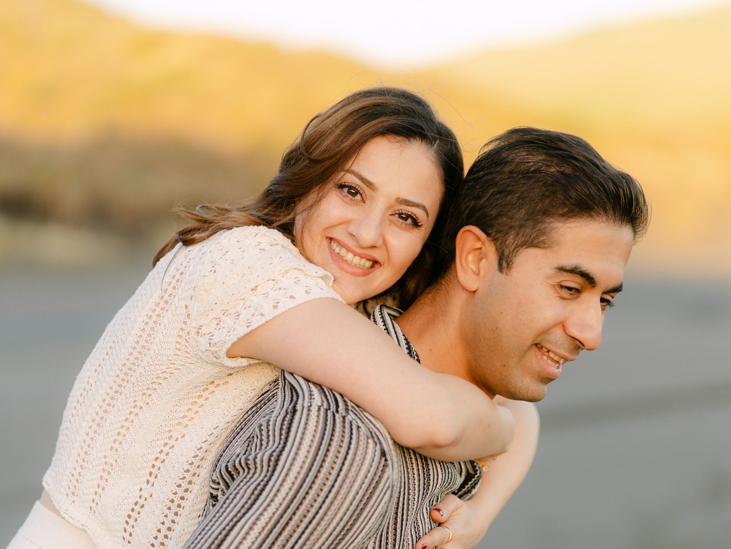 A smiling woman hugs a man from behind outdoors during golden hour at a beach.