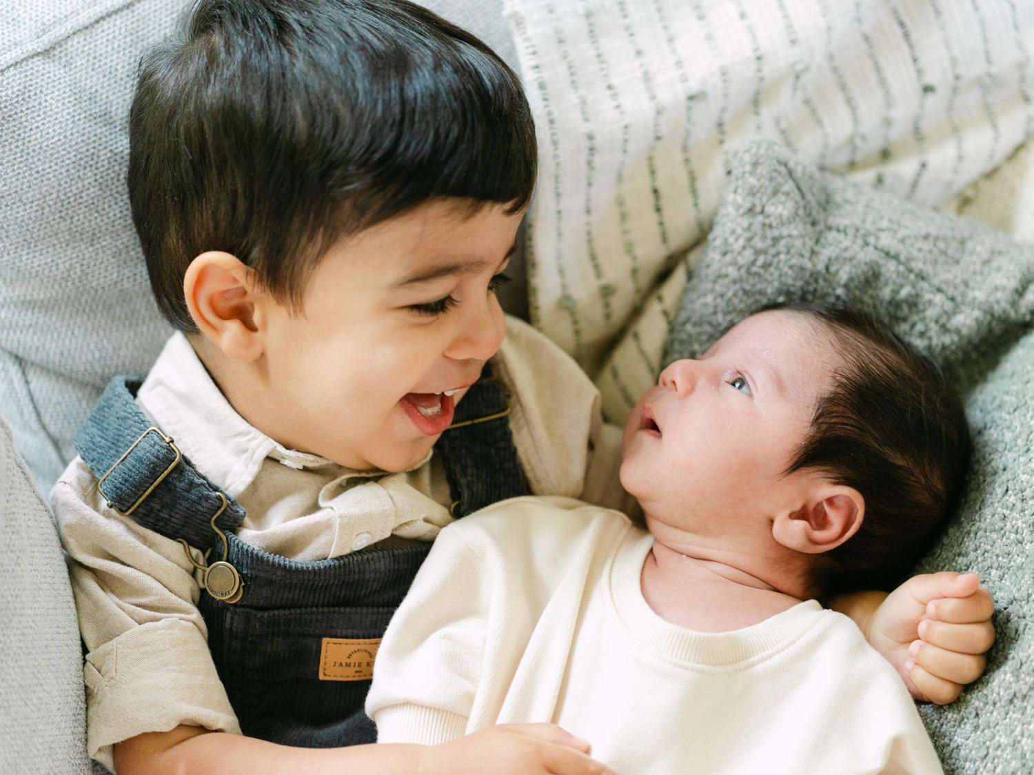A toddler boy in denim overalls smiles at a newborn baby lying on a soft blanket.