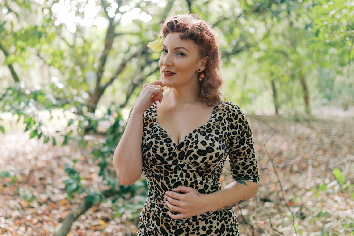 Woman in leopard print dress with yellow flower hair accessory posing outdoors among green trees.