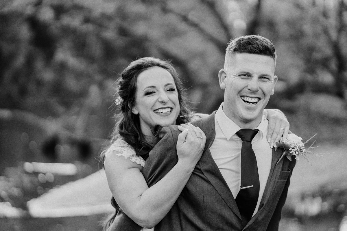 Black and white photo of a smiling bride and groom in formal wedding attire posing outdoors.
