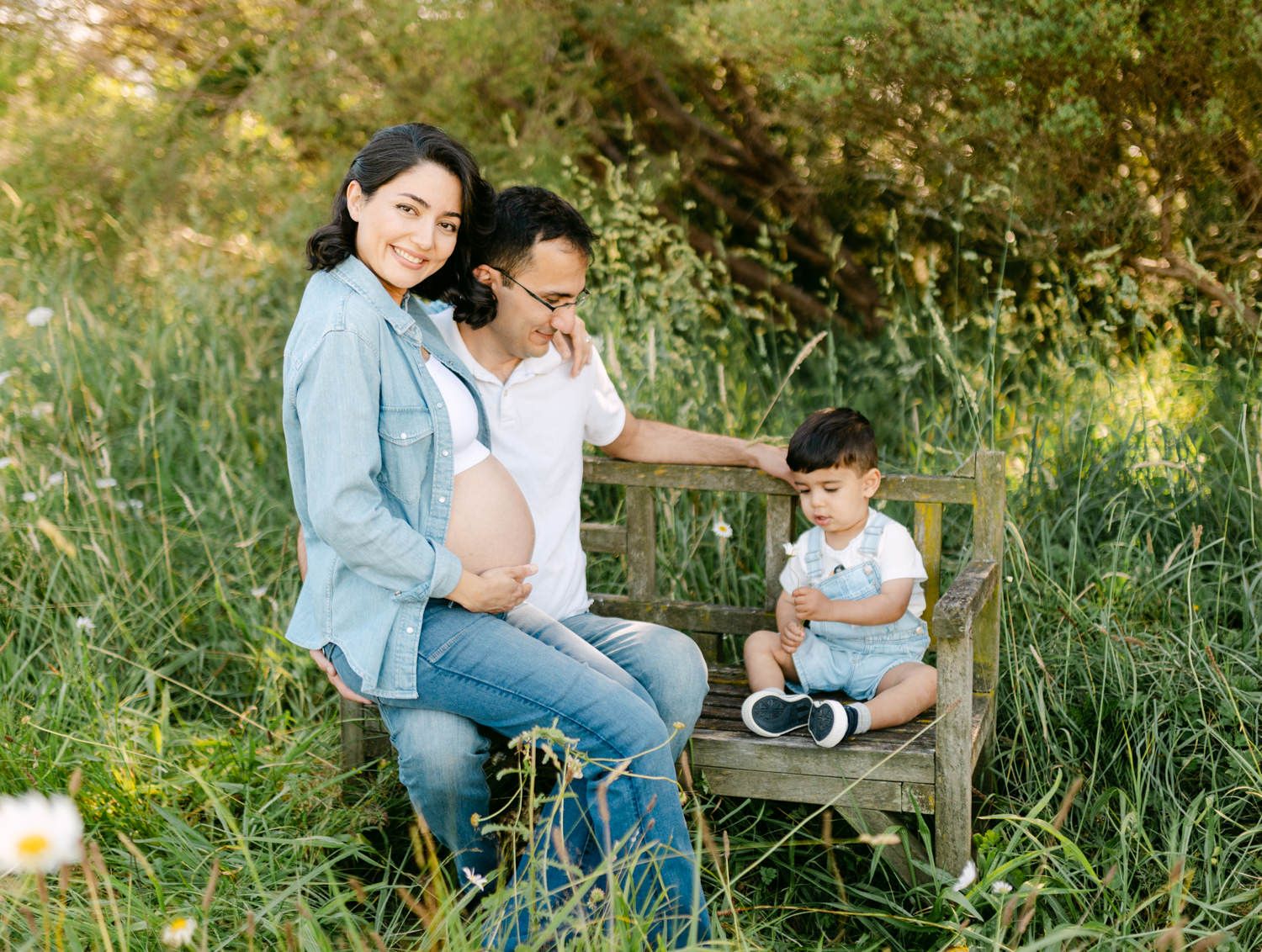 Pregnant woman with partner and toddler son posing on wooden bench in lush green meadow for maternity photo.