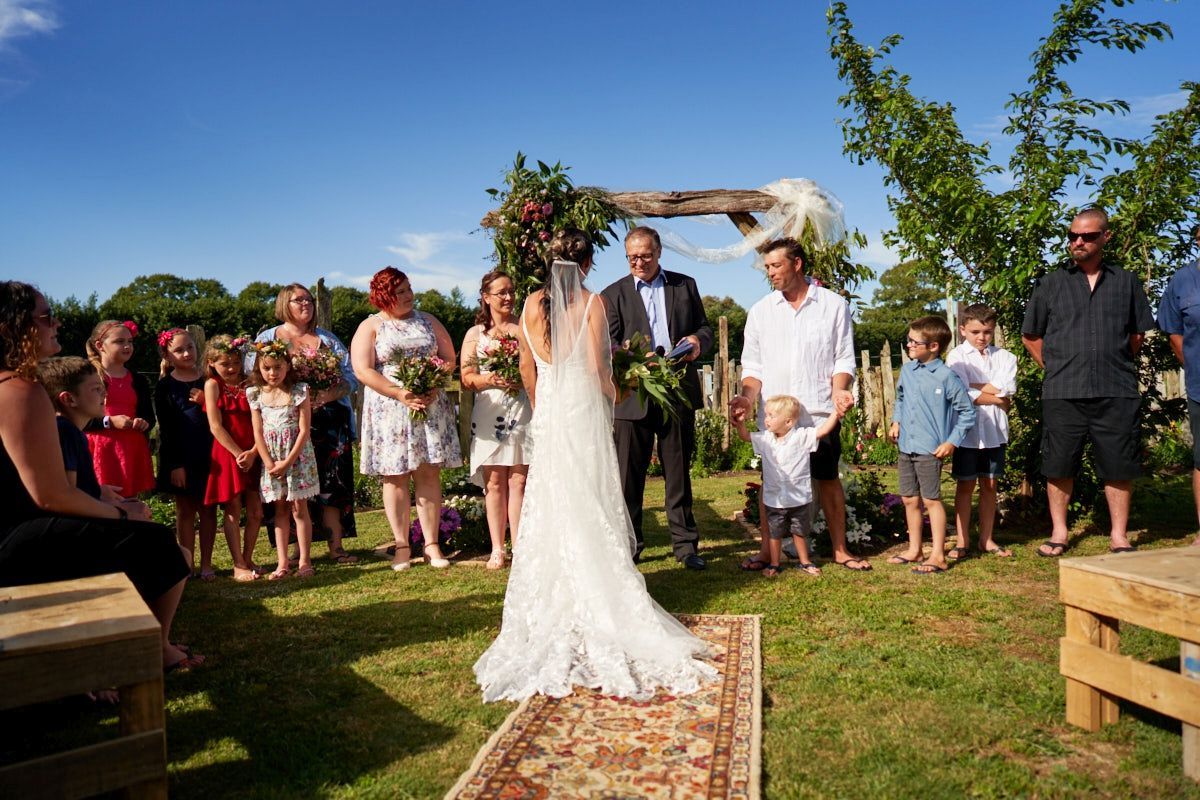 Outdoor wedding ceremony on green lawn with bride in lace gown, groom, officiant, and guests under floral arch on sunny day.