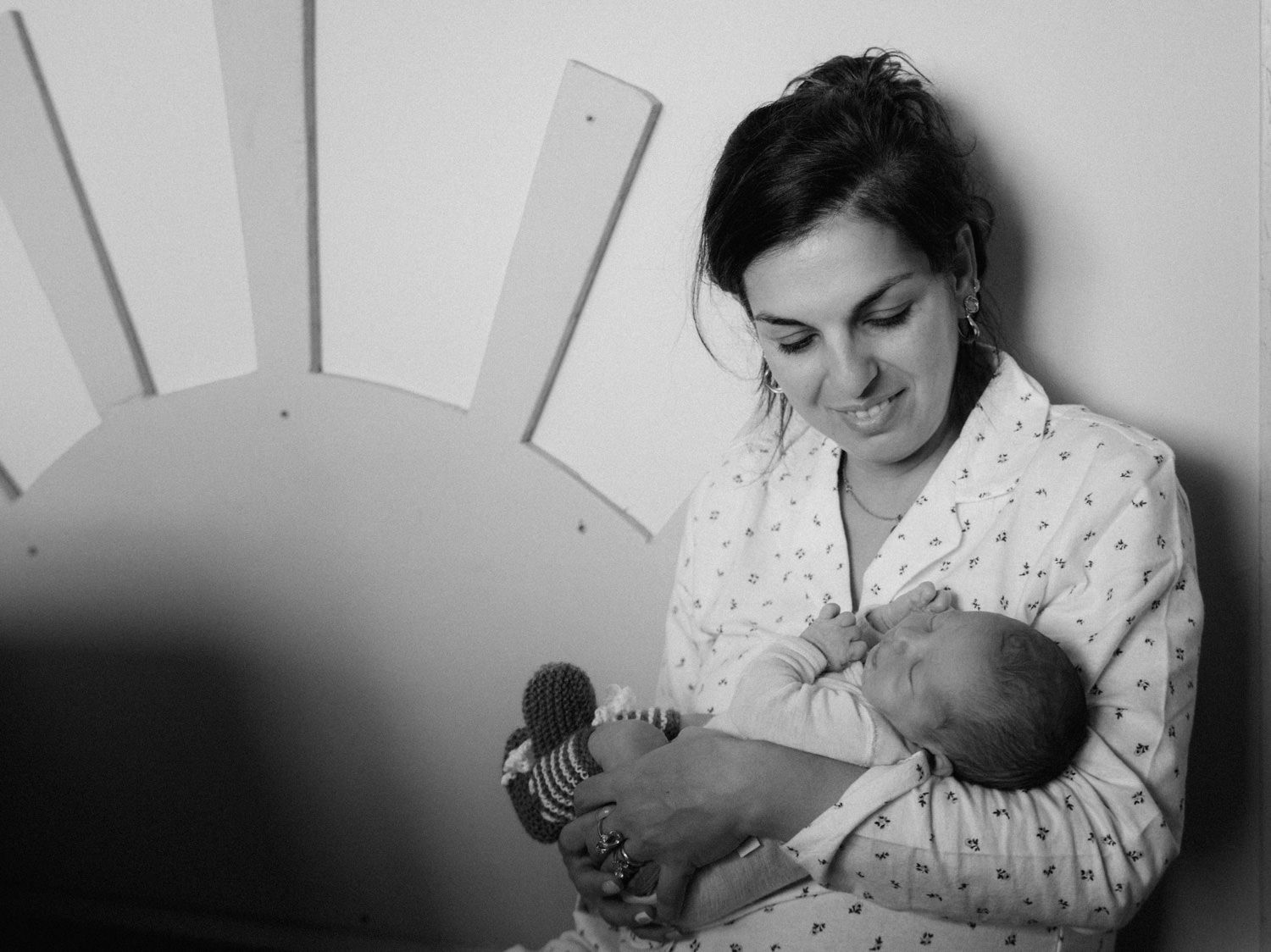 Black and white photo of a mother tenderly holding her newborn baby against a nursery wall with sun decor.