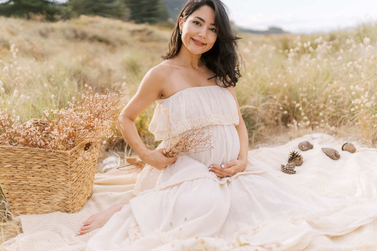Pregnant woman in white strapless dress sitting in a field with a wicker basket, cradling her baby bump.