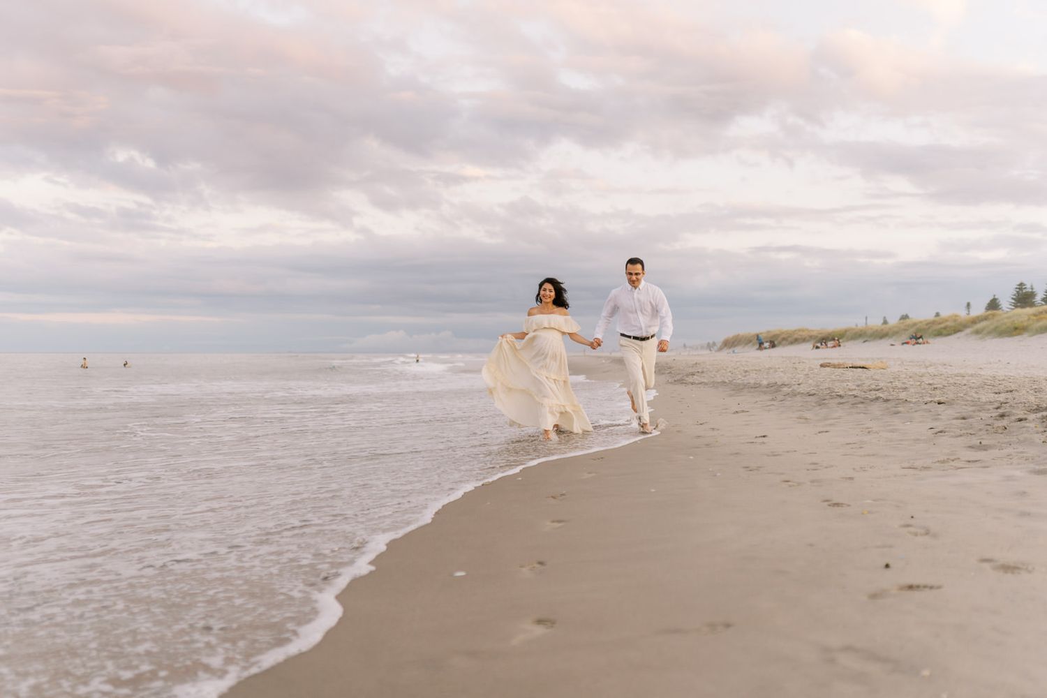 Couple holding hands running along a sandy beach at sunset, woman in flowing white dress, waves gently lapping the shore.