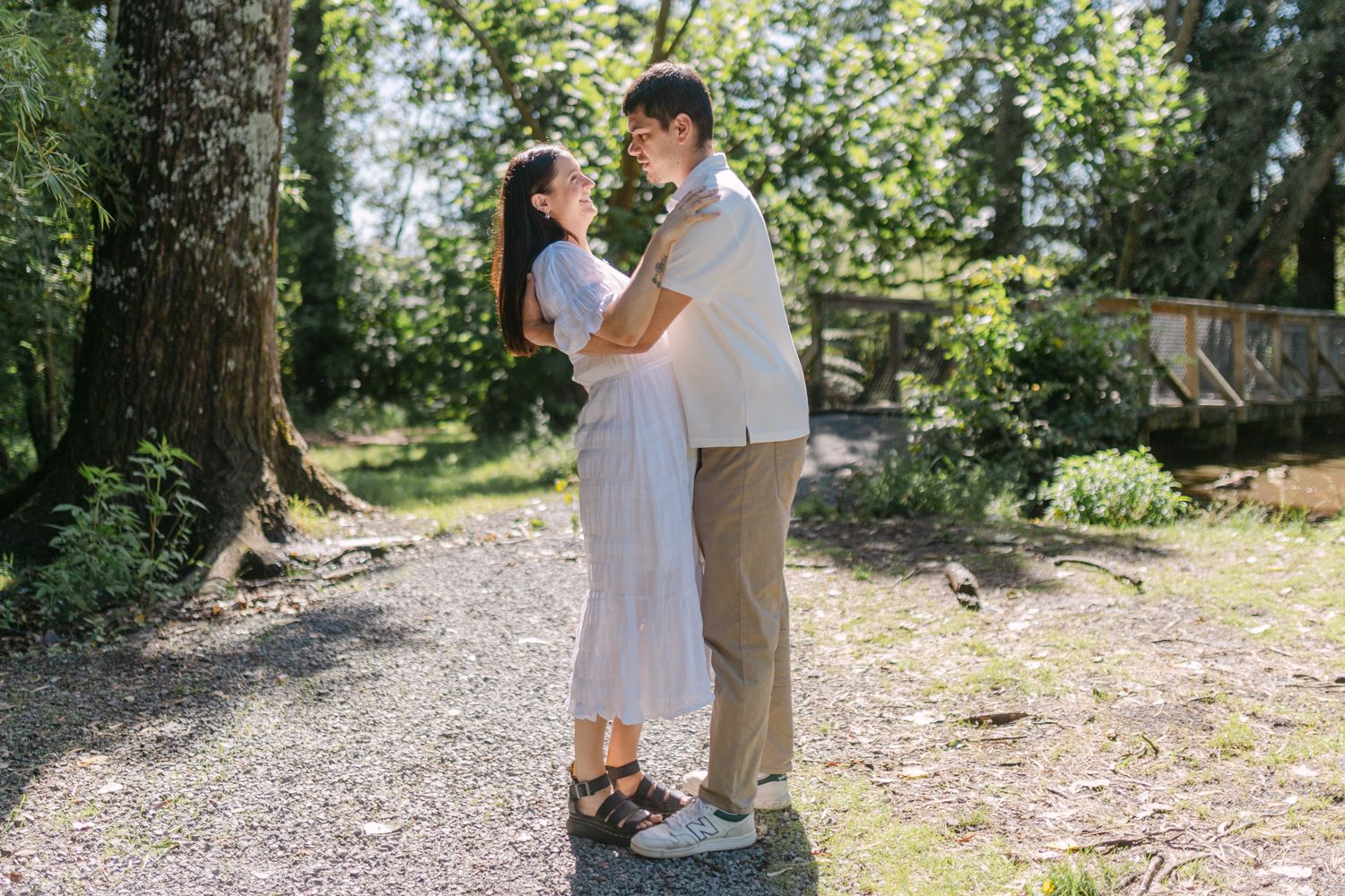 Couple embracing and gazing at each other in a sunlit garden surrounded by lush green trees.