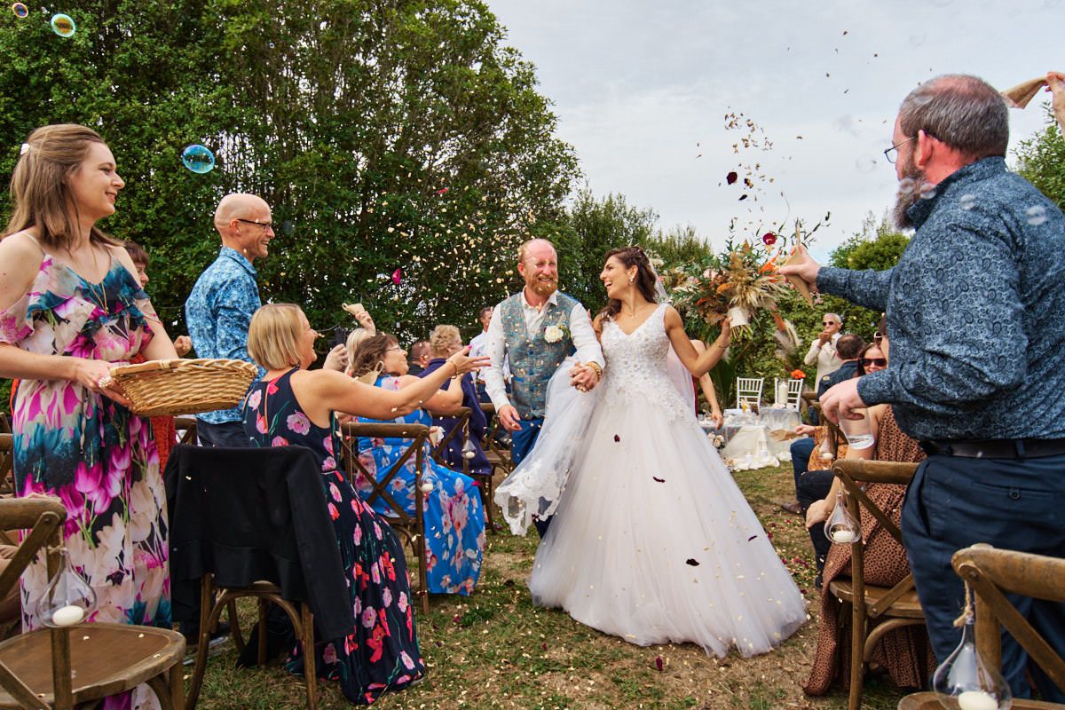 Bride and groom walk through confetti shower surrounded by smiling wedding guests outdoors.