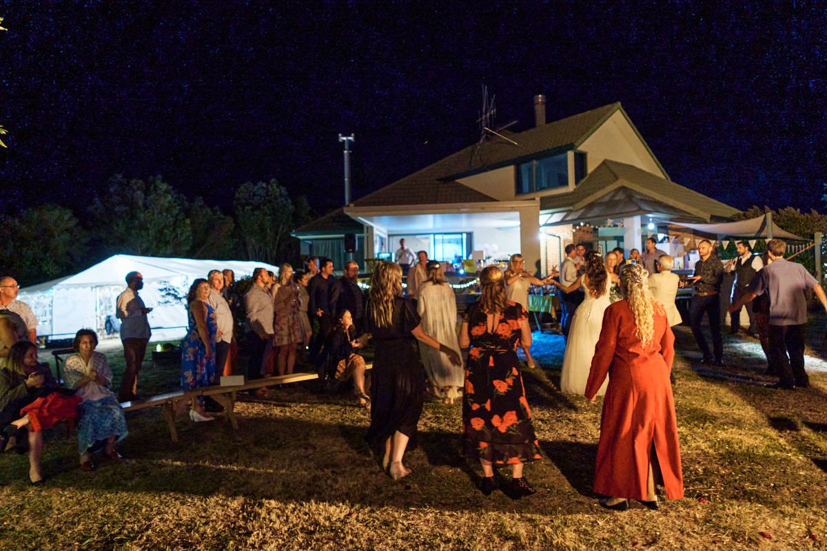 Outdoor evening gathering with crowd of people socializing near illuminated white tent and house at night event.