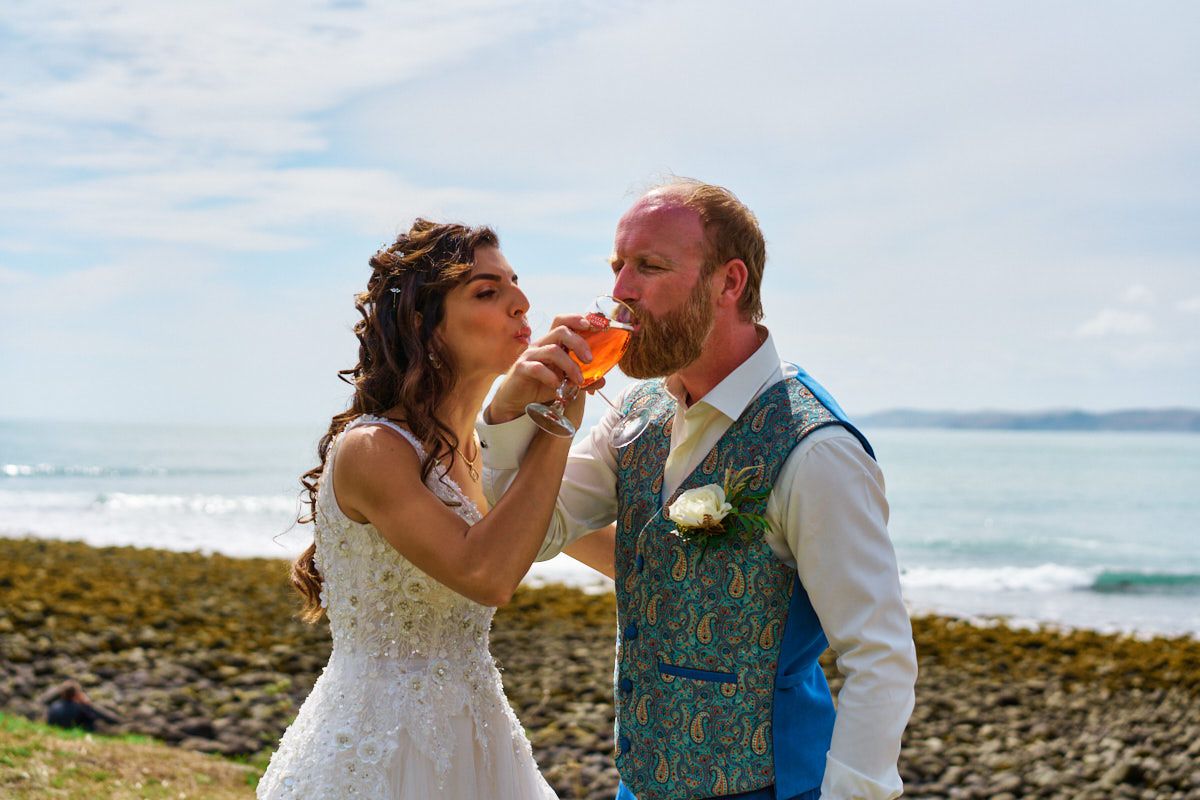 Bride and groom sharing a drink by the ocean on their wedding day, with scenic coastal views in the background.