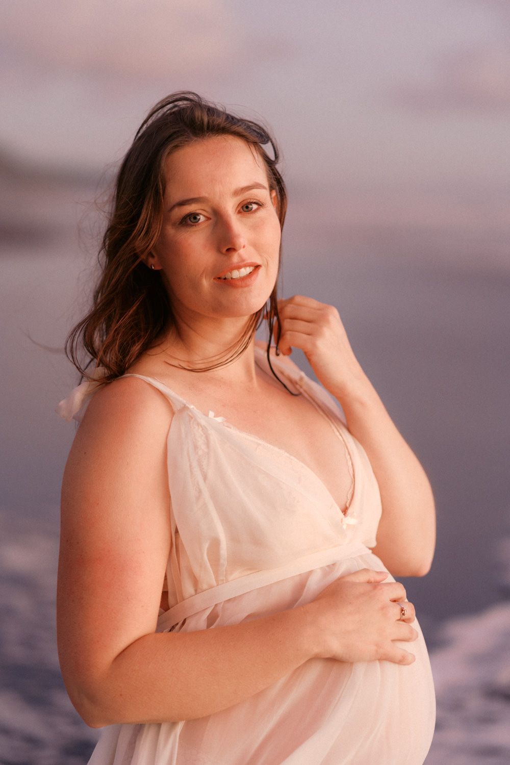 Pregnant woman in sheer white dress poses gracefully at beach during golden hour sunset.
