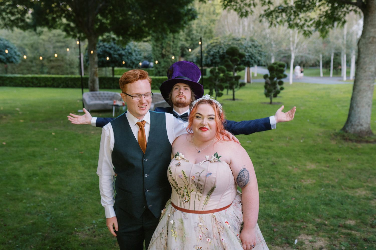 Bride in floral gown with two grooms, one in purple hat, posing outdoors at a garden wedding celebration.