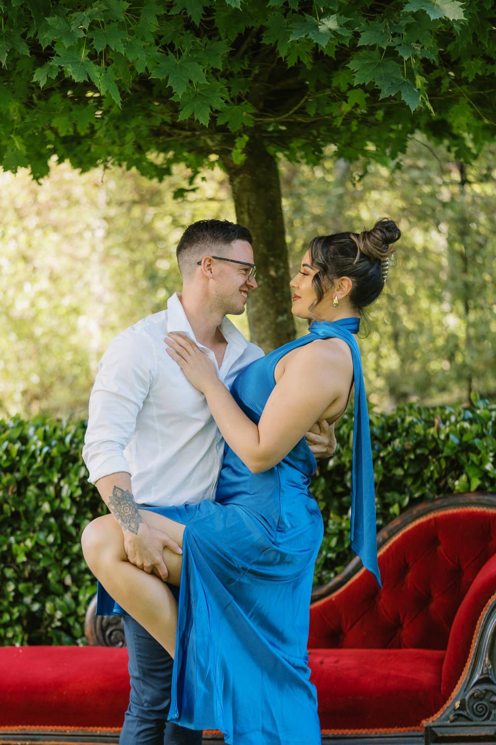 Couple embracing on a red velvet sofa outdoors, man in white shirt and woman in blue halter dress.