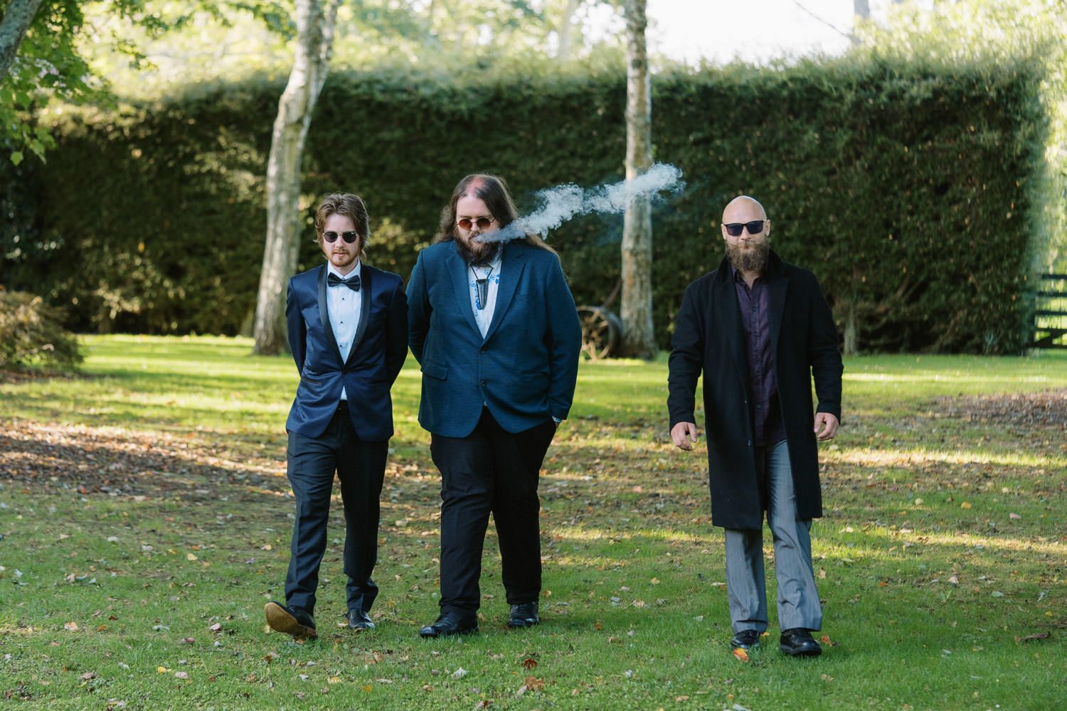 Three men in formal suits walking outdoors on green grass with trees in the background.