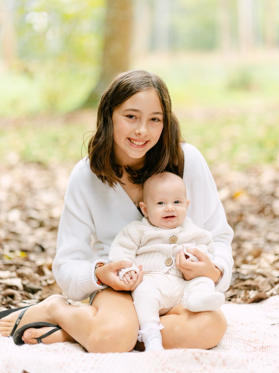 A smiling teenage girl sits cross-legged on a blanket outdoors, cradling a happy baby in a cream knit outfit.