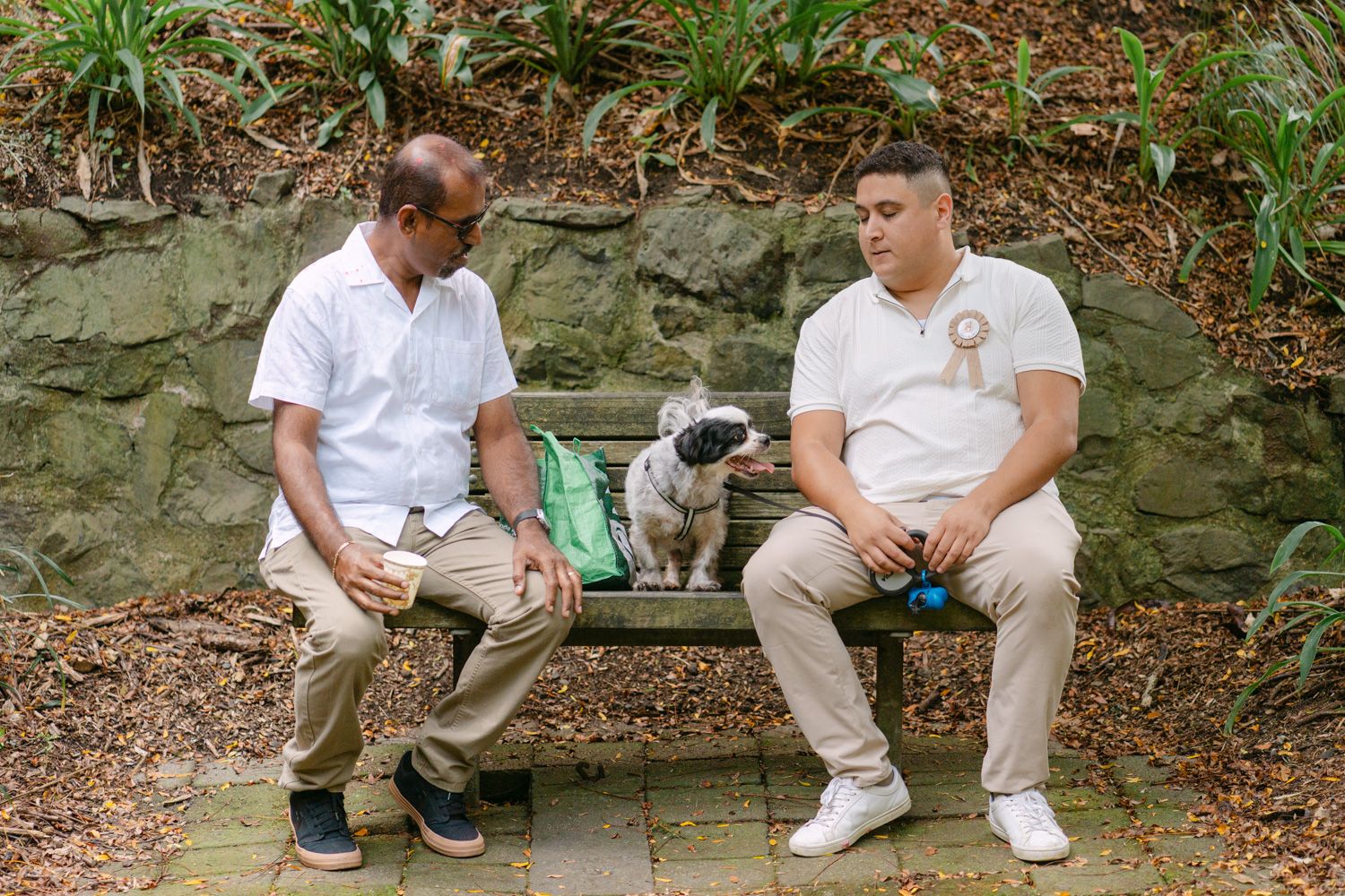 Two men sit on a bench outdoors with a pug dog between them, surrounded by lush greenery.
