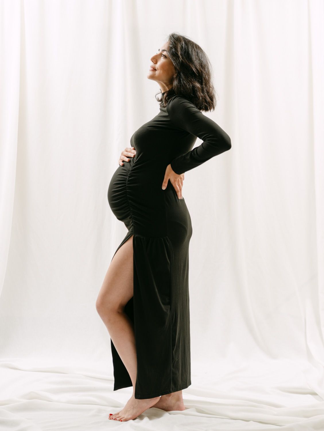 Pregnant woman in black long-sleeve maxi dress with slit, cradling baby bump, posing against white curtain backdrop.