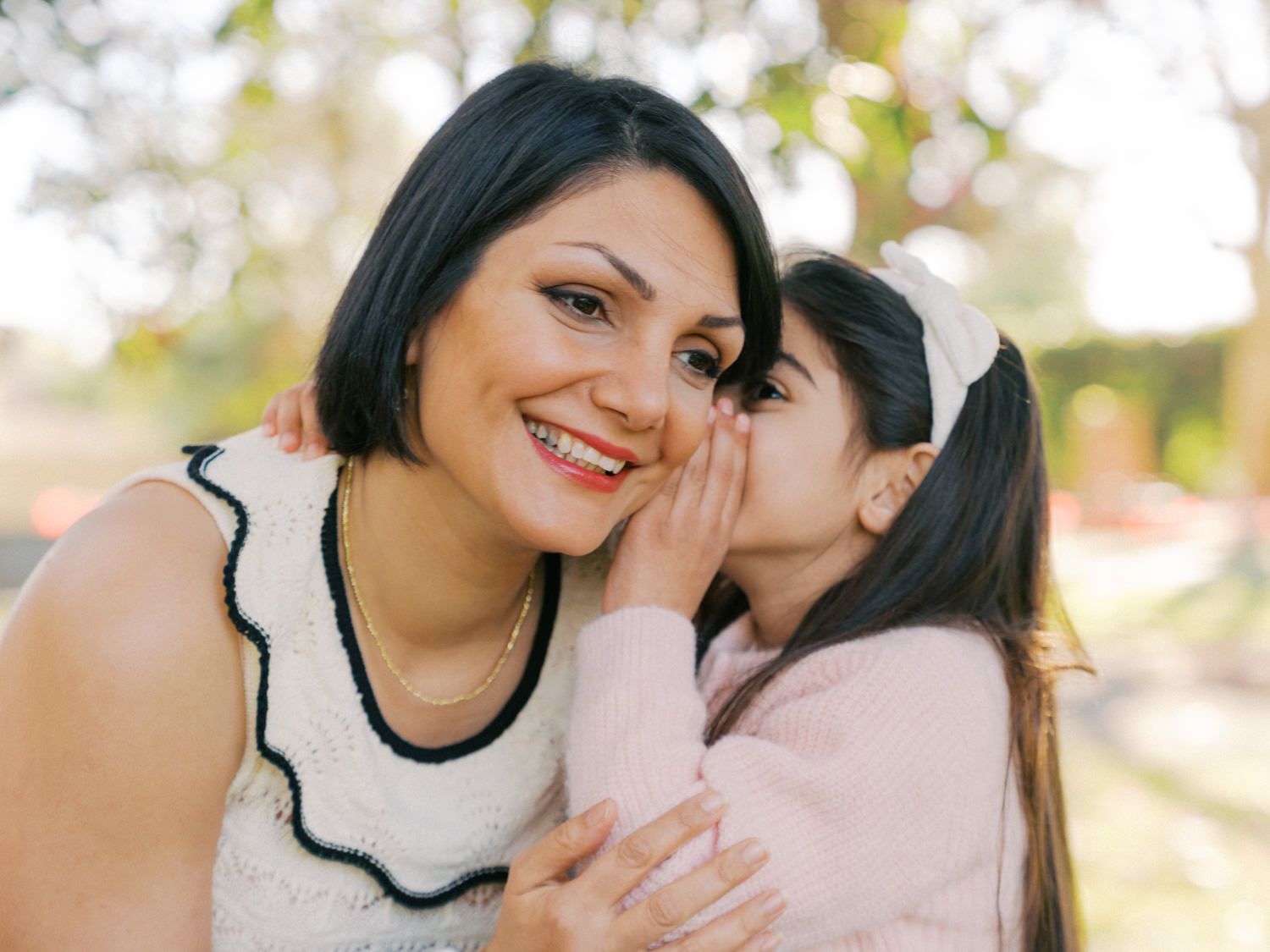 A young girl whispers in her smiling mother's ear outdoors in a warm, sunlit park setting.
