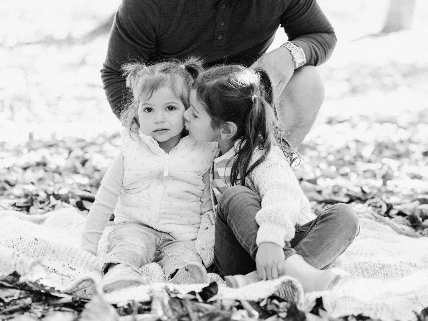 Black and white photo of two young sisters sitting on a blanket outdoors, sharing a sweet moment together.