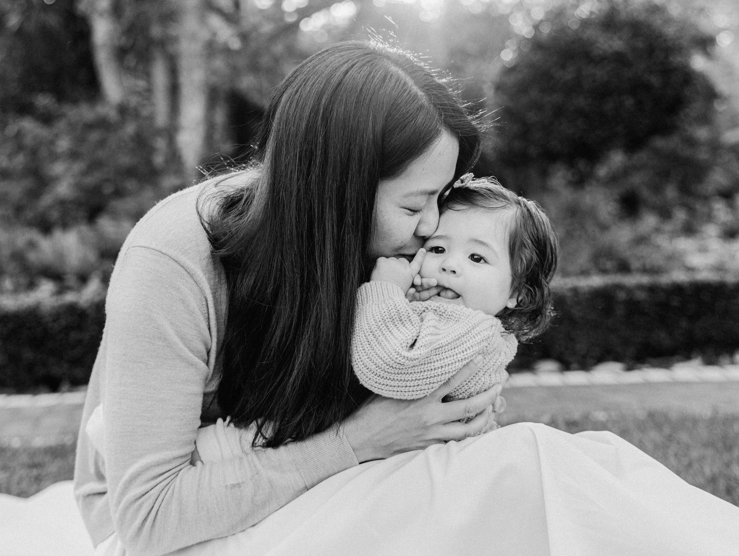 Black and white photo of a mother tenderly kissing her smiling toddler outdoors in a garden setting.