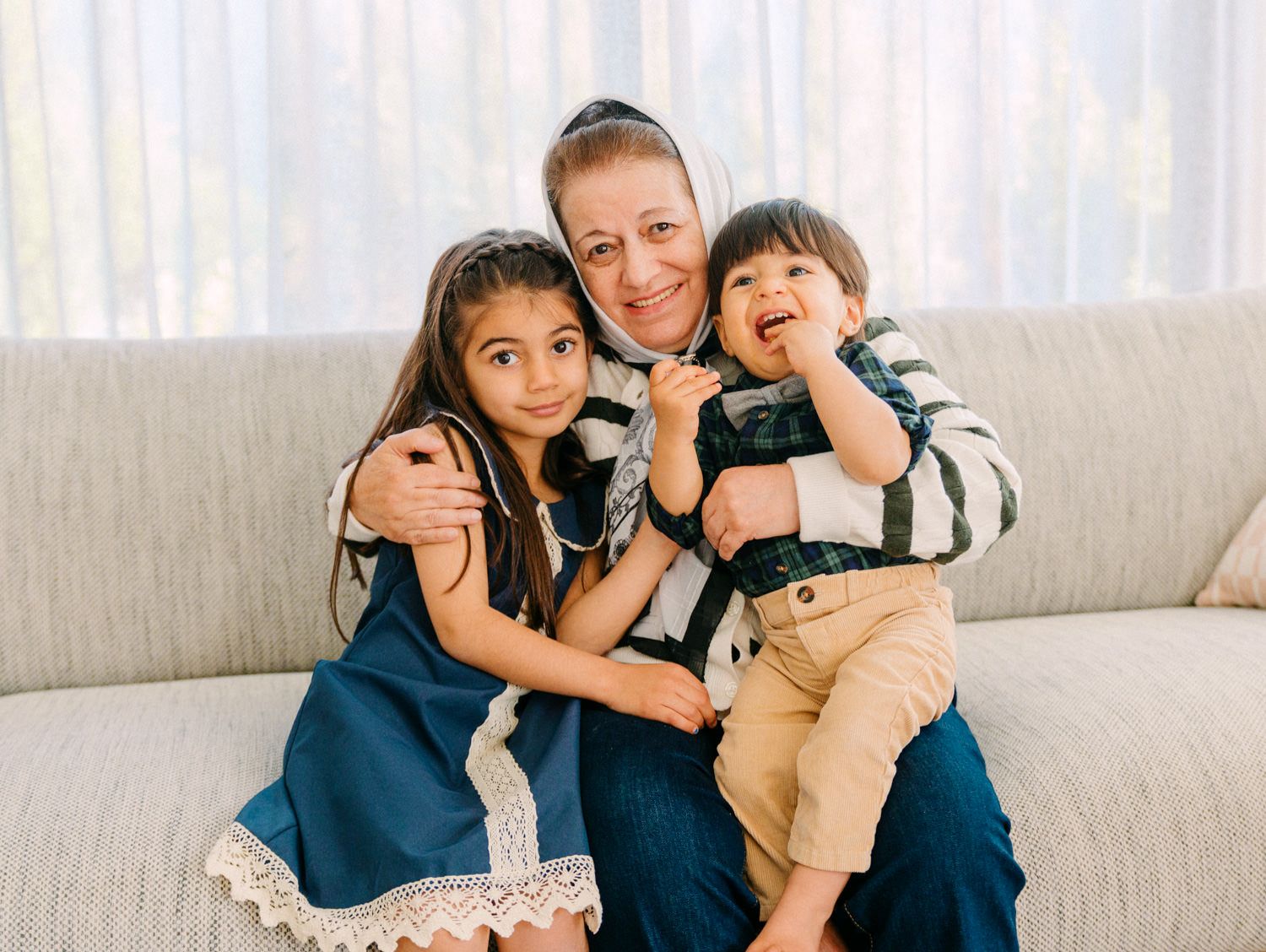 Smiling adult with two young children sitting together on a beige sofa in a bright living room.
