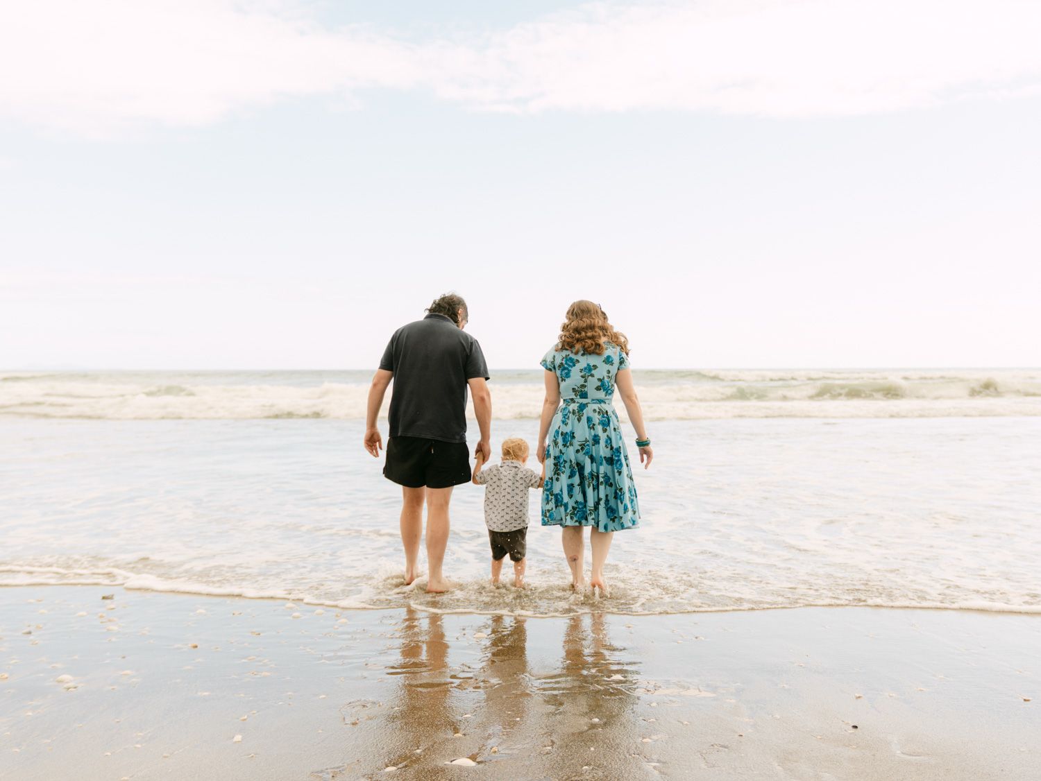 Family of three holding hands at the ocean's edge on a sandy beach, viewed from behind.