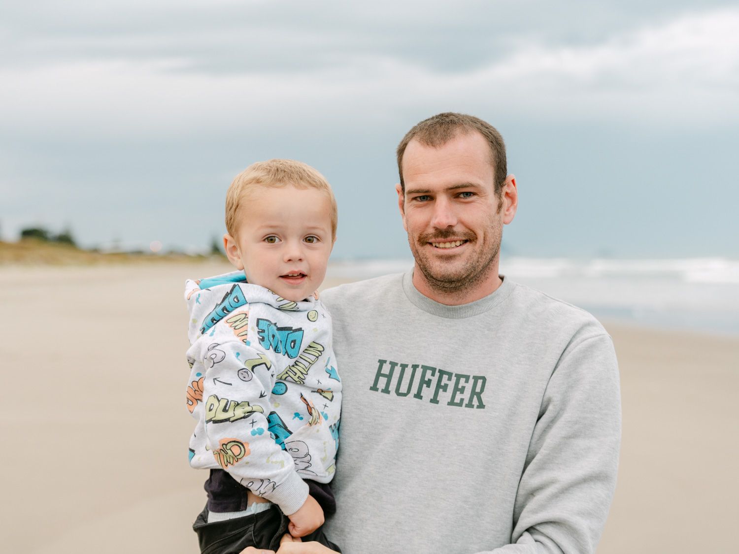 Man in Huffer sweatshirt holding a young toddler on a sandy beach with cloudy skies.