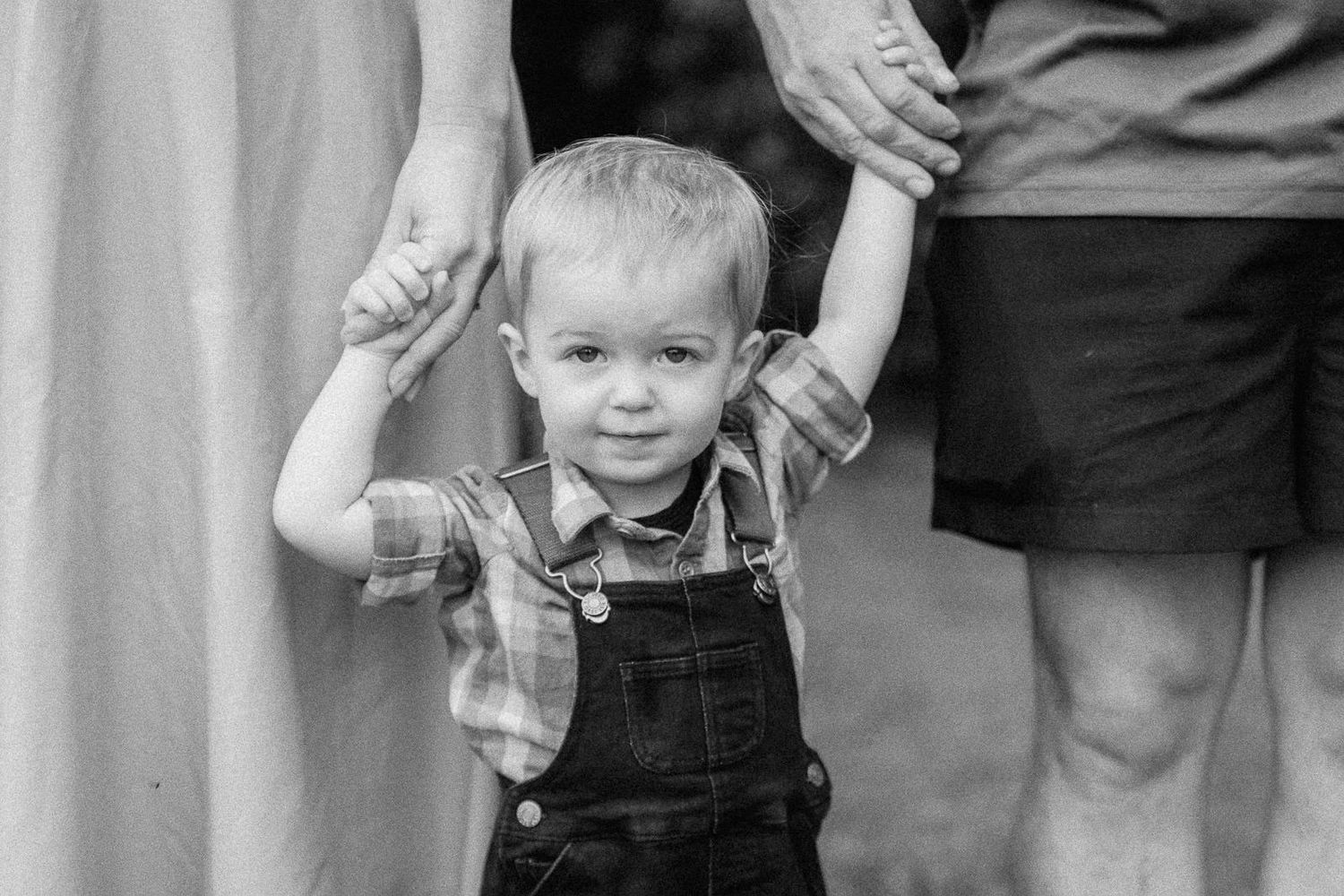 Toddler boy in overalls and plaid shirt holds hands with parents in black and white family portrait.