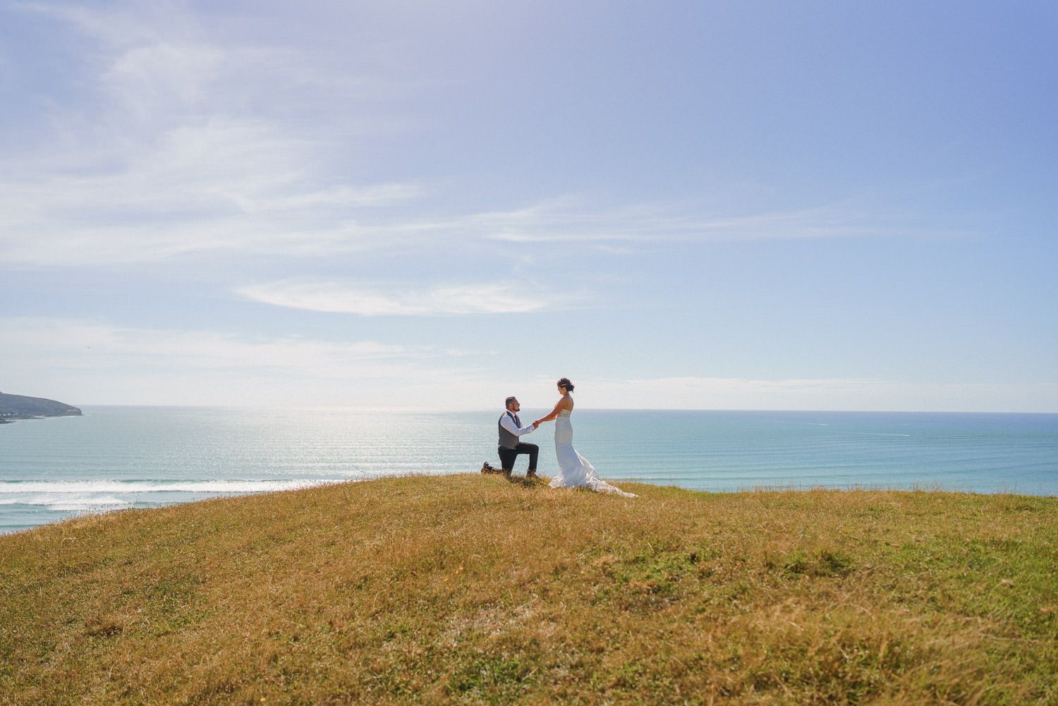 Man proposes to woman on grassy coastal cliff overlooking ocean under blue sky.