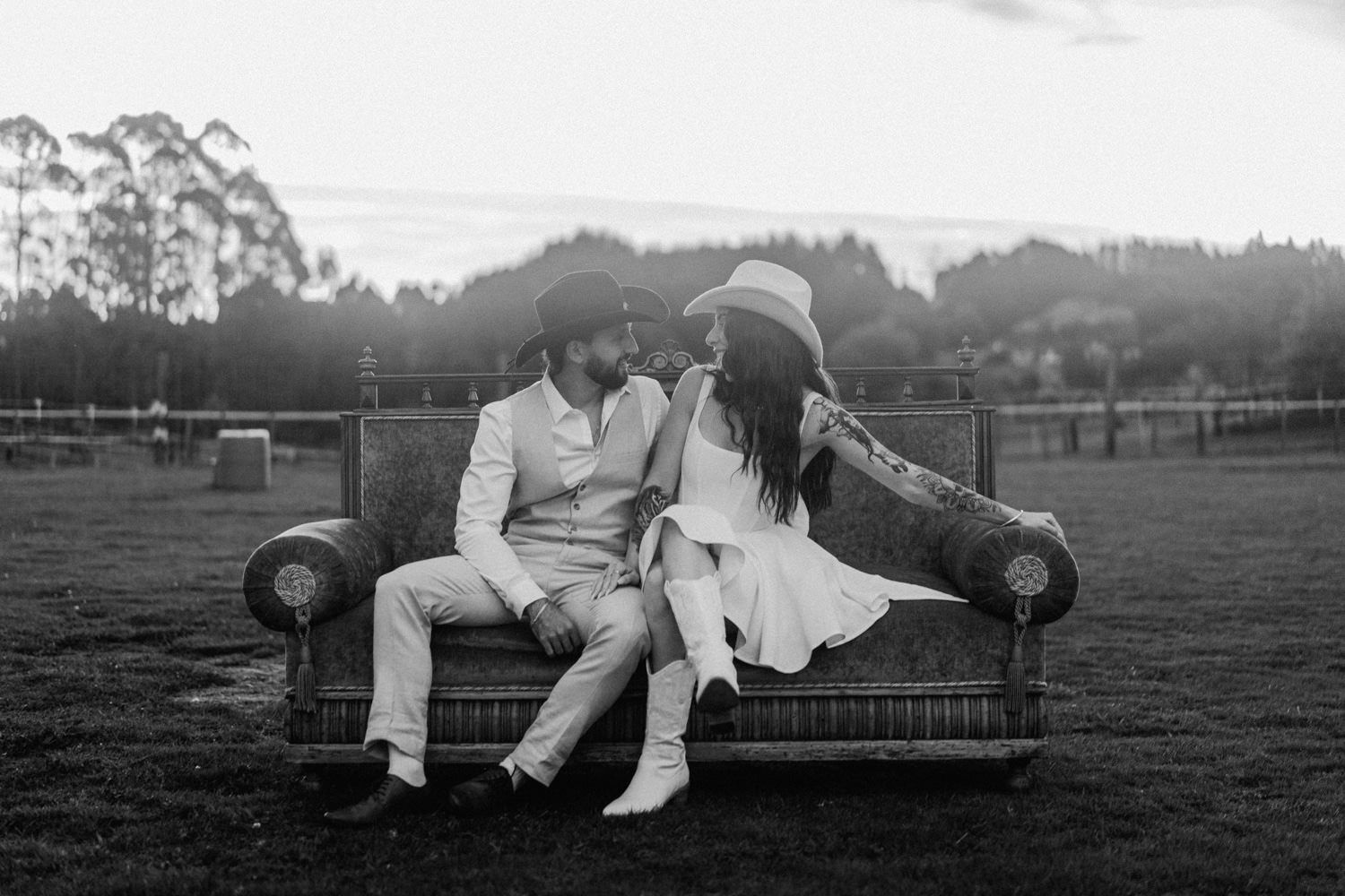 Black and white photo of a couple sitting closely on a vintage sofa outdoors in a rustic field setting.