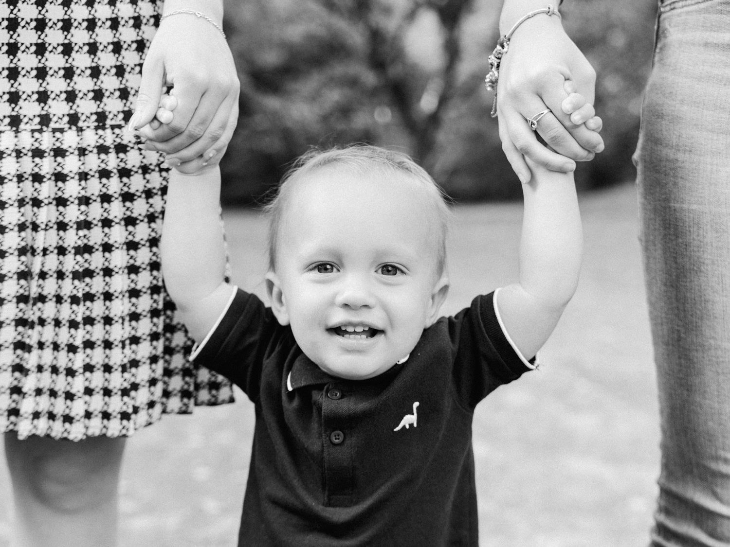 Black and white photo of a smiling toddler boy holding hands with two adults outdoors.