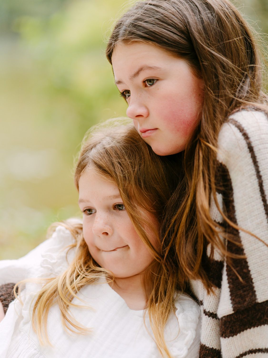 Two young girls with long brown hair pose together outdoors, the older girl embracing the younger in a tender sibling portrait.