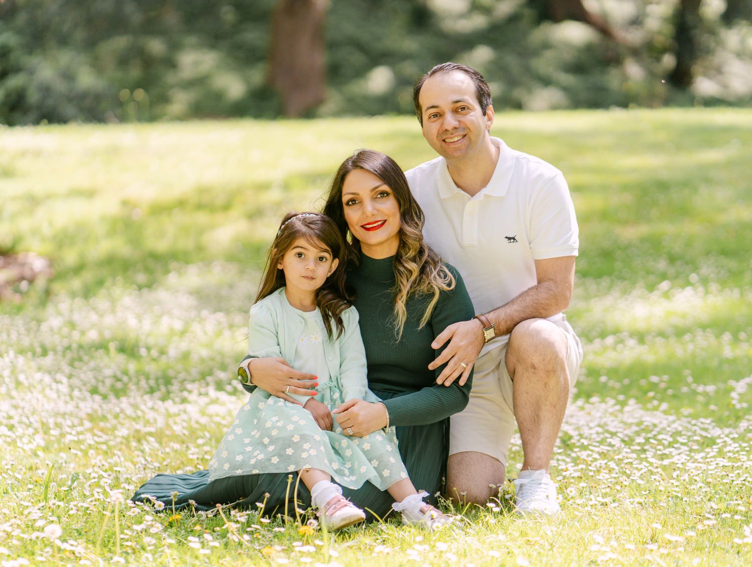 Family of three smiling together in a sunny park with wildflowers, mother, father, and young daughter posing outdoors.