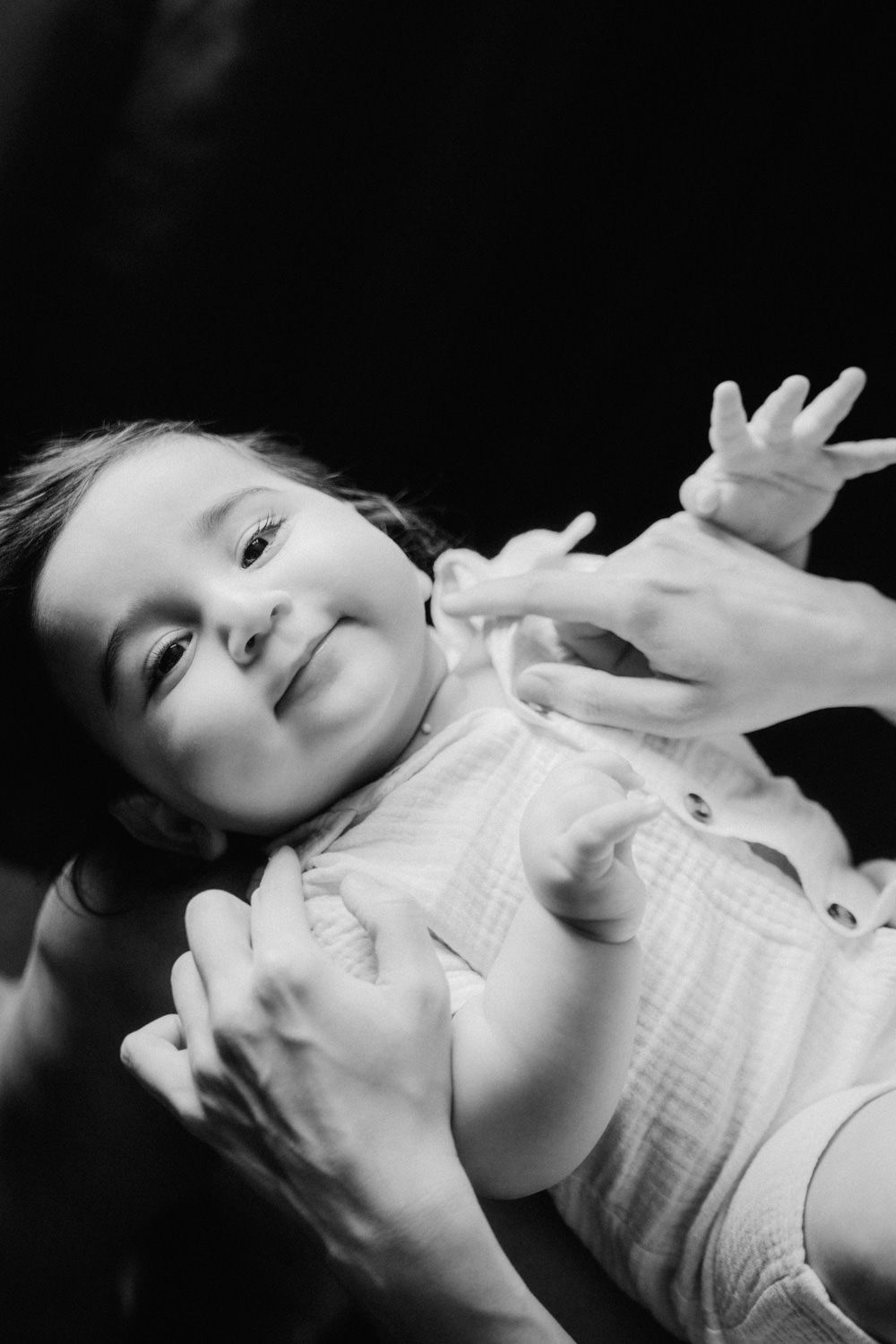 Black and white photo of a smiling baby being held by adult hands against a dark background.