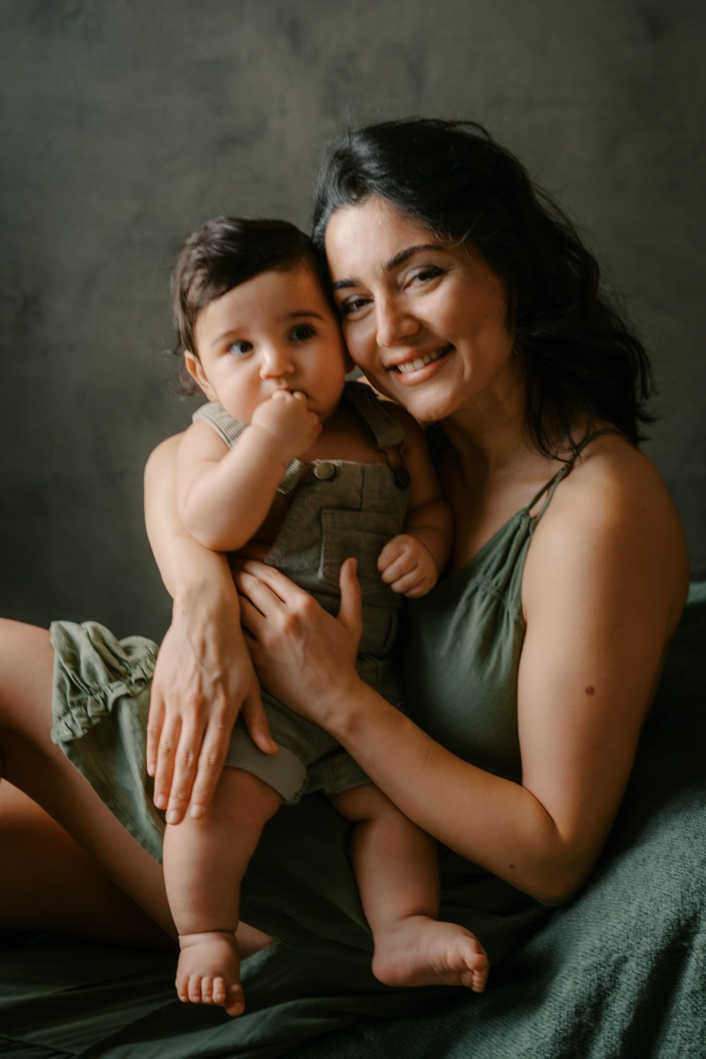 Smiling woman in olive dress holding a cute baby in green overalls against a dark studio background.