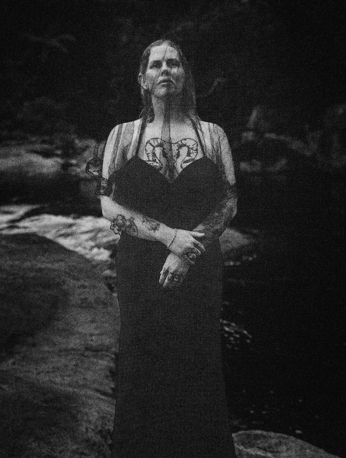 Black and white photo of a tattooed woman in a dark dress standing by a rocky river, looking upward.