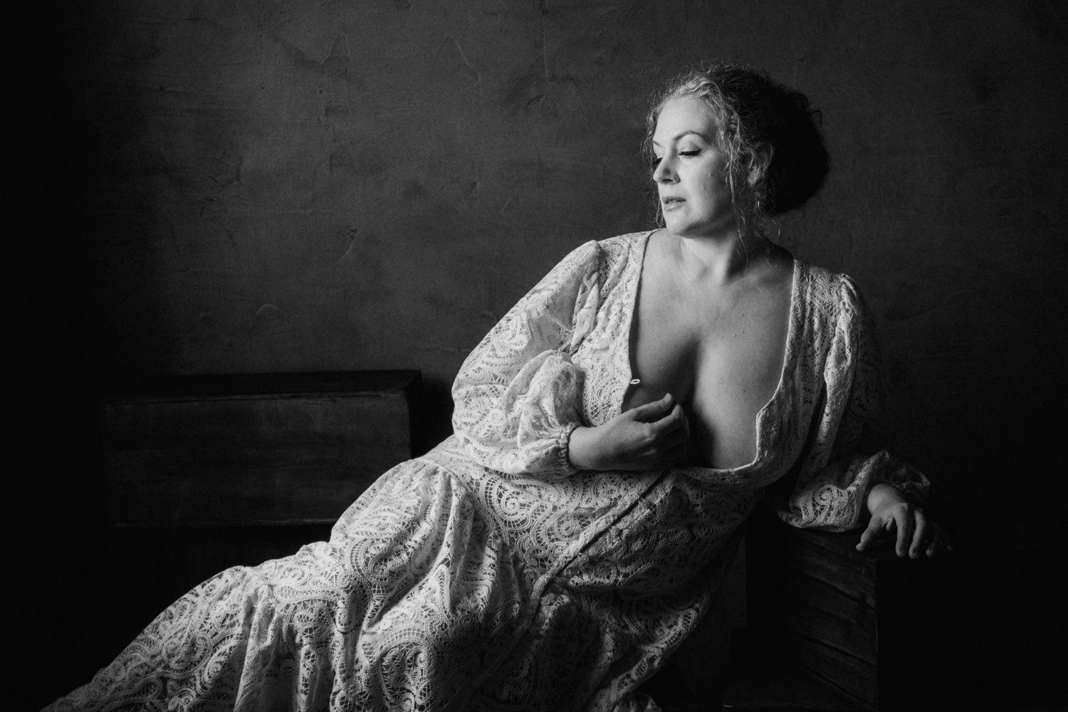 Black and white portrait of a woman in lace robe seated elegantly, looking away in soft dramatic lighting.
