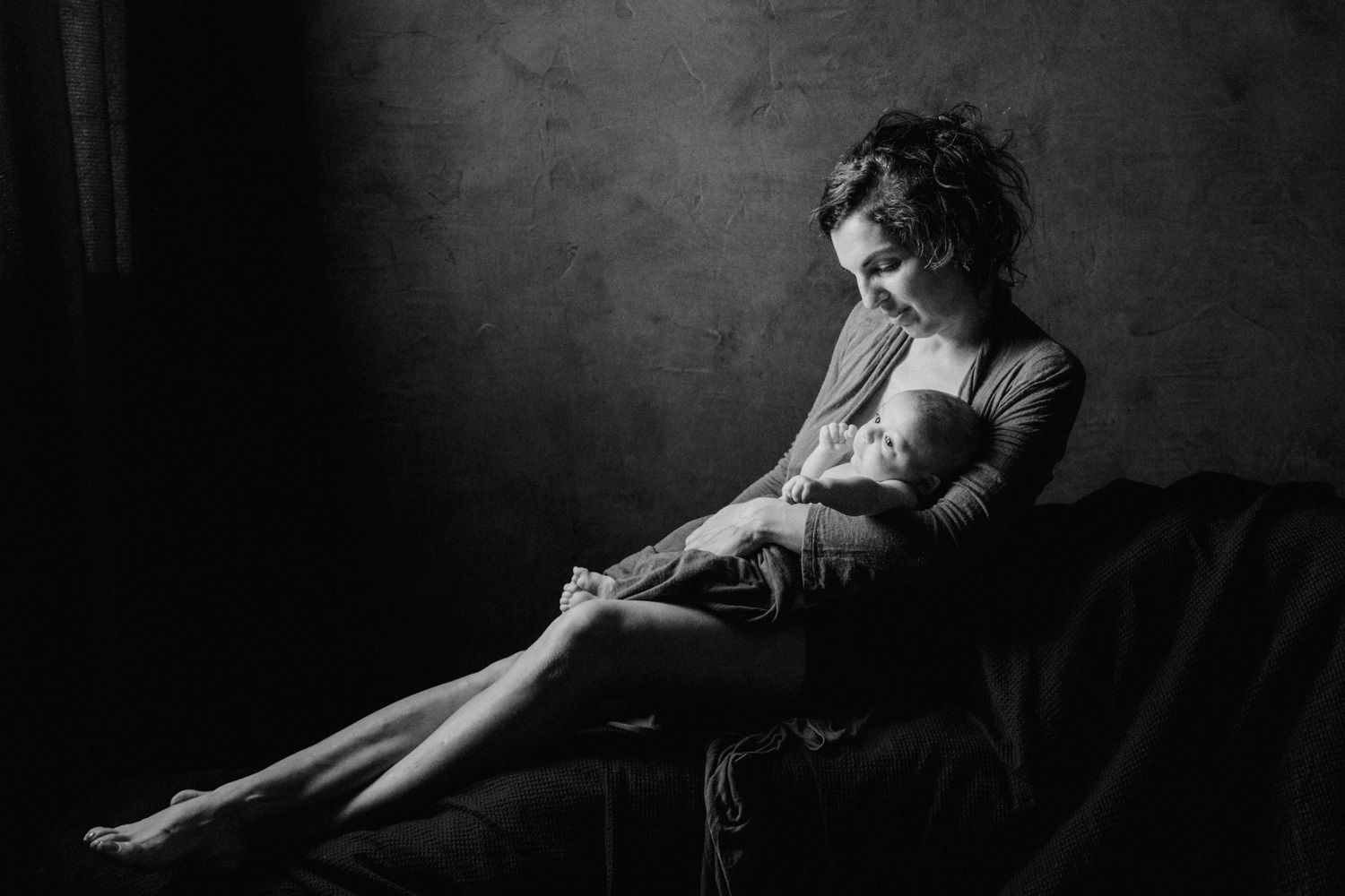 Black and white photo of a mother tenderly breastfeeding her newborn baby while seated in soft natural light.