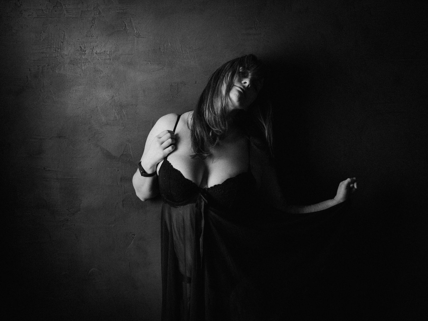 Black and white artistic portrait of a woman in dark lingerie posing dramatically against a textured dark wall.